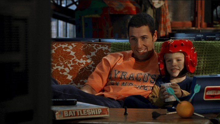 Adam Sandler abrazando a un niño con casco, ambos viendo la televisión en una escena de Big Daddy.