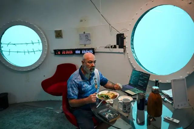 Koch está sentado en su escritorio, comiendo un plato de comida, el cual se encuentra al lado de un computador portátil. Detrás de Koch, dos ventanas redondas dan a las profundidades. En medio de ellas, hay un dibujo de Koch, y un reloj que marca la hora, además del tiempo que lleva bajo el agua. 