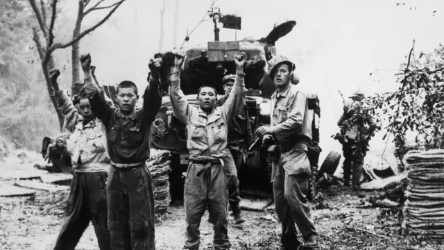 Tres hombres coreanos con uniforme militar levantan las manos, con lo que parece ser un soldado estadounidense armado detrás de ellos.