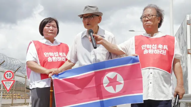 Ahn Hak-sop sostiene una bandera de Corea del Norte con dos voluntarios con chalecos rojos parados a su lado
