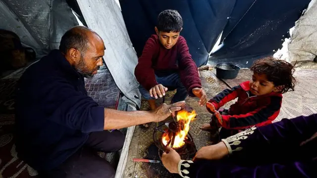 Un padre, una madre y sus dos hijos pequeños descalzos intentan calentar sus manos junto a un pequeño fuego