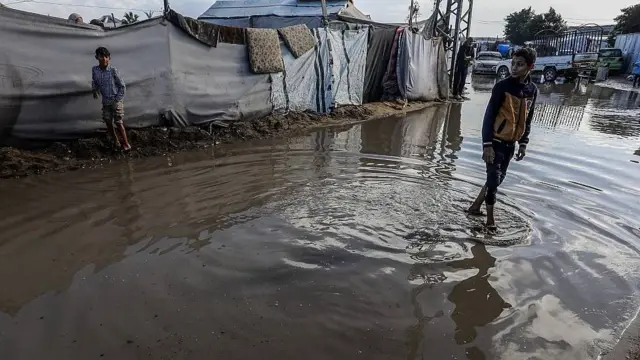 Colchones mojados colgados en carpas precarias, mientras un niño camina con el agua hasta los tobillos