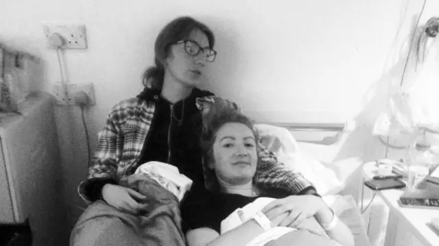 Fotografía en blanco y negro de Lowri en el hospital en septiembre de 2015, esperando unas pruebas junto a su hermana. Lowri está acostada en la cama y su hermana la abraza. Ambas llevan el pelo recogido. La hermana de Lowri lleva una sudadera con capucha.