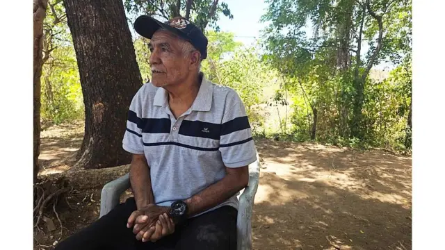 Don Lorenzo sentado en una silla de plástico. Al fondo se ven árboles. Su rostro denota desesperanza